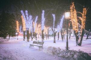 invierno parque a noche con Navidad decoraciones, brillante linternas, pavimento cubierto con nieve y arboles Clásico película estético. foto