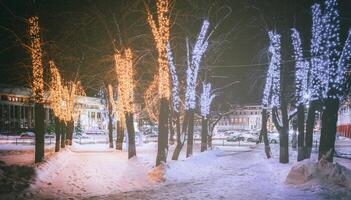 invierno parque a noche con Navidad decoraciones, brillante linternas, pavimento cubierto con nieve y arboles Clásico película estético. foto