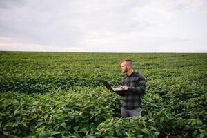 joven agrónomo sostiene tableta toque almohadilla computadora en el soja campo y examinando cultivos antes de cosecha. agronegocios concepto. agrícola ingeniero en pie en un soja campo con un tableta en verano. foto