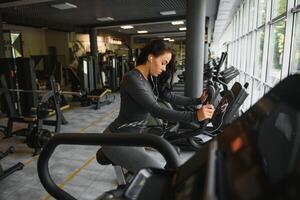 Young woman with headphones doing exercises on stationery bicycle in a gym or fitness center. Young sporty woman in gym listen music from smartphone. Women doing cardio exercises photo
