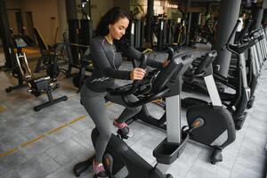 joven mujer con auriculares haciendo ejercicios en papelería bicicleta en un gimnasio o aptitud centro. joven deportivo mujer en gimnasio escucha música desde teléfono inteligente mujer haciendo cardio ejercicios foto