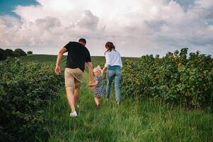 Happy family walking in the park. Mom, dad and daughter walk outdoors, parents holding the baby girl's hands. Childhood, parenthood, family bonds, marriage concept. photo