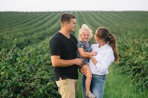 el hija abrazando padres en naturaleza. mamá, papá y niña niño pequeño, caminar en el césped. contento joven familia gasto hora juntos, afuera, en vacaciones, al aire libre. el concepto de familia fiesta foto