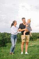 el hija abrazando padres en naturaleza. mamá, papá y niña niño pequeño, caminar en el césped. contento joven familia gasto hora juntos, afuera, en vacaciones, al aire libre. el concepto de familia fiesta foto