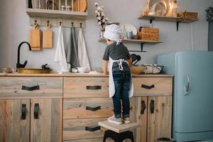 joven contento mamá y su bebé cocinar galletas a hogar en el cocina. Navidad hecho en casa pan de jengibre. linda chico con madre en blanco uniforme y sombrero cocido chocolate galletas. foto