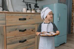 joven contento mamá y su bebé cocinar galletas a hogar en el cocina. Navidad hecho en casa pan de jengibre. linda chico con madre en blanco uniforme y sombrero cocido chocolate galletas foto