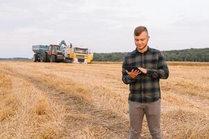 Young attractive farmer with laptop standing in wheat field with combine harvester in background. photo