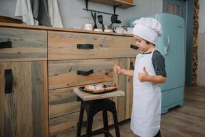 joven contento mamá y su bebé cocinar galletas a hogar en el cocina. Navidad hecho en casa pan de jengibre. linda chico con madre en blanco uniforme y sombrero cocido chocolate galletas foto