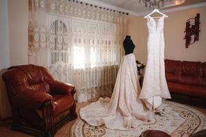 Beautiful bride's white wedding dress hangs near the bed in a hotel room with flowers at the bottom. Bridal morning before ceremony photo