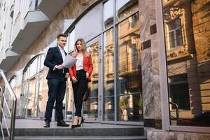 Two confident business colleagues walking on office terrace. Mid adult business man and woman discussing document outdoors on coffee break. Working during break concept. photo