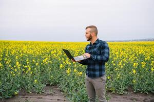 Young agronomist holds laptop in rape field. Agribusiness concept. agricultural engineer standing in a rape field with a laptop in summer. photo