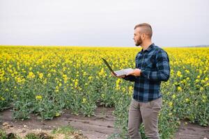 Young agronomist holds laptop in rape field. Agribusiness concept. agricultural engineer standing in a rape field with a laptop in summer. photo