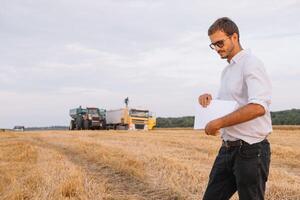joven agrónomo hombre en pie en trigo campo comprobación calidad mientras combinar segador trabajando foto