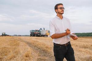 Young agronomist man standing on wheat field checking quality while combine harvester working photo