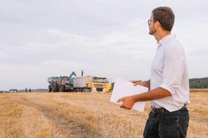 joven agrónomo hombre en pie en trigo campo comprobación calidad mientras combinar segador trabajando foto