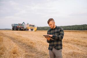 Young agronomist man standing on wheat field checking quality while combine harvester working photo