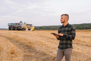 Happy young farmer engineer with notebook standing on wheat field while combine harvester working in background. photo