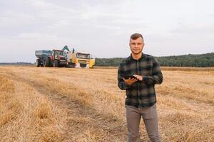 Young agronomist man standing on wheat field checking quality while combine harvester working photo