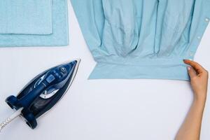 Female hand ironing clothes top view isolated on white background. Young woman with iron ironing man's shirt seen from above during housework. Blue iron on white table photo