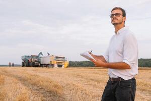 joven atractivo granjero con ordenador portátil en pie en trigo campo con combinar segador en antecedentes. foto