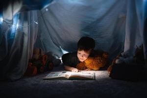 niño chico leyendo con libro y Linterna y osito de peluche oso en carpa. antes de yendo a cama. foto