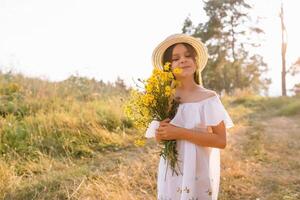 al aire libre Disparo de agradable mirando joven niña con bronceado sano piel, vestido en blanco vestir y verano sombrero, poses en parque con confidente satisfecho expresión, gustos recreación. hermosa joven mujer. foto