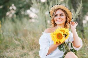 Outdoor shot of pleasant looking female with tanned healthy skin, dressed in white dress and summer hat, poses in park with confident satisfied expression, likes recreation. Beautiful young woman. photo