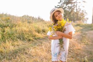 Outdoor shot of pleasant looking young girl with tanned healthy skin, dressed in white dress and summer hat, poses in park with confident satisfied expression, likes recreation. Beautiful young woman. photo