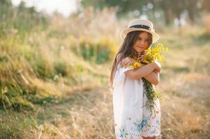 Outdoor shot of pleasant looking young girl with tanned healthy skin, dressed in white dress and summer hat, poses in park with confident satisfied expression, likes recreation. Beautiful young woman. photo