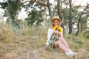 Outdoor shot of pleasant looking female with tanned healthy skin, dressed in white dress and summer hat, poses in park with confident satisfied expression, likes recreation. Beautiful young woman photo