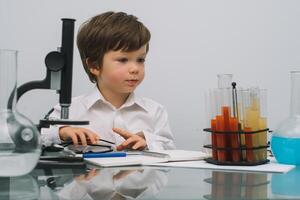 el chico con un microscopio y varios vistoso matraces en un blanco antecedentes. un chico haciendo experimentos en el laboratorio. explosión en el laboratorio. Ciencias y educación foto