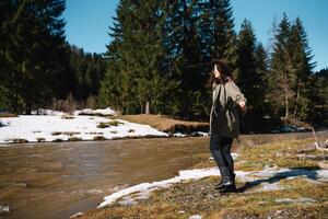 Stylish girl traveling in woods. Young woman in hat exploring in summer forest, looking at river. Travel and wanderlust concept. Space for text. photo