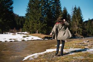 Stylish girl traveling in woods. Young woman in hat exploring in summer forest, looking at river. Travel and wanderlust concept. Space for text. photo