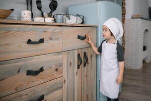 joven chico linda en el cocina cocinar cocinero en blanco uniforme y sombrero cerca mesa. Navidad hecho en casa pan de jengibre. el chico cocido el chocolate galletas. foto