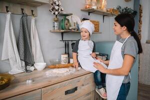 joven contento mamá y su bebé cocinar galletas a hogar en el cocina. Navidad hecho en casa pan de jengibre. linda chico con madre en blanco uniforme y sombrero cocido chocolate galletas. foto