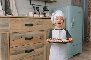 joven chico linda en el cocina cocinar cocinero en blanco uniforme y sombrero cerca mesa. Navidad hecho en casa pan de jengibre. el chico cocido el chocolate galletas foto