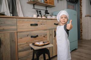 joven chico linda en el cocina cocinar cocinero en blanco uniforme y sombrero cerca mesa. Navidad hecho en casa pan de jengibre. el chico cocido el chocolate galletas foto
