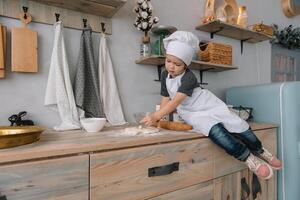 joven chico linda en el cocina cocinar cocinero en blanco uniforme y sombrero cerca mesa. Navidad hecho en casa pan de jengibre. el chico cocido el chocolate galletas foto