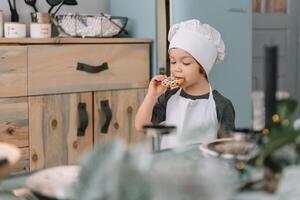 joven chico linda en el cocina cocinar cocinero en blanco uniforme y sombrero cerca mesa. Navidad hecho en casa pan de jengibre. el chico cocido el chocolate galletas. foto