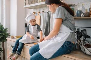 Young happy mom and her baby cook cookies at home in the kitchen. Christmas Homemade Gingerbread. cute boy with mother in white uniform and hat cooked chocolate cookies photo