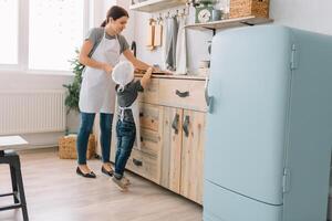 joven contento mamá y su bebé cocinar galletas a hogar en el cocina. Navidad hecho en casa pan de jengibre. linda chico con madre en blanco uniforme y sombrero cocido chocolate galletas foto