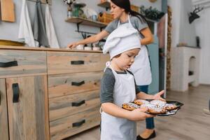 Young happy mom and her baby cook cookies at home in the kitchen. Christmas Homemade Gingerbread. cute boy with mother in white uniform and hat cooked chocolate cookies photo