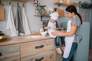 joven contento mamá y su bebé cocinar galletas a hogar en el cocina. Navidad hecho en casa pan de jengibre. linda chico con madre en blanco uniforme y sombrero cocido chocolate galletas foto