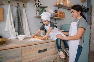 joven contento mamá y su bebé cocinar galletas a hogar en el cocina. Navidad hecho en casa pan de jengibre. linda chico con madre en blanco uniforme y sombrero cocido chocolate galletas foto