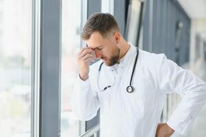 Sad doctor leaning against the wall in hospital corridor. photo