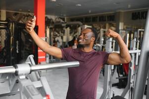 smiling young sportsman showing biceps and taking selfie with smartphone in gym. photo