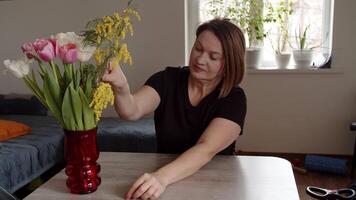 midden- oud vrouw zetten bloemen in een vaas in de keuken, mimosa's en tulpen. video