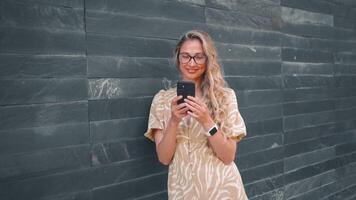 sorridente donna utilizzando smartphone pendente su grigio parete nel città video