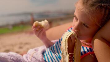 mignonne peu fille en mangeant banane et pain sur sablonneux plage près rivage video