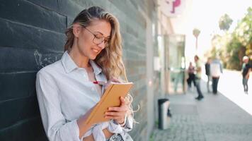 a woman in glasses leaning on a stone wall is holding a notbook and writing on it video